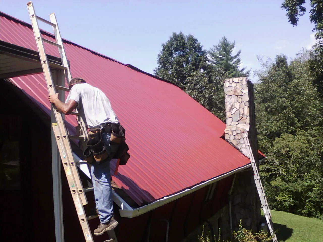 Roofer on a ladder beside a red metal roof and stone chimney — Soffit & Fascia Repair by Roofing Brothers in Heathsville